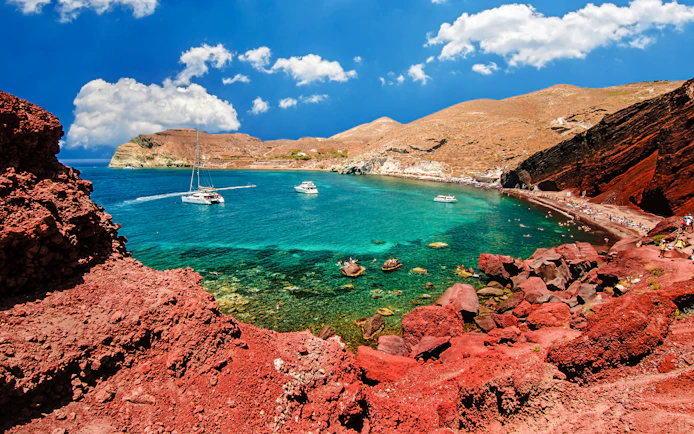 Catamarans on turquoise waters near Red Beach, Santorini, with red cliffs and tourists on the shore.