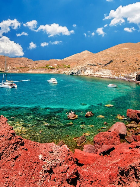 Catamarans on turquoise waters near Red Beach, Santorini, with red cliffs and tourists on the shore.