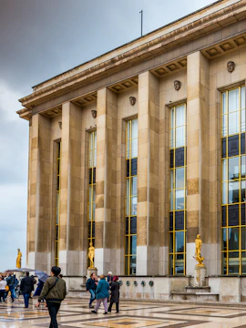 Visitors walking outside Musée de l'Homme, Paris, with golden statues and tall columns.