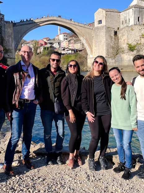 Group of tourists by the Neretva River with Mostar Bridge in Bosnia in the background.