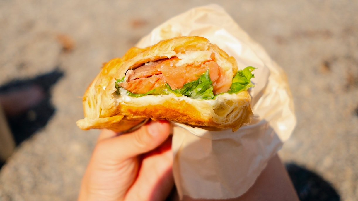 Hand holding a croissant sandwich with salmon and lettuce on a beach.