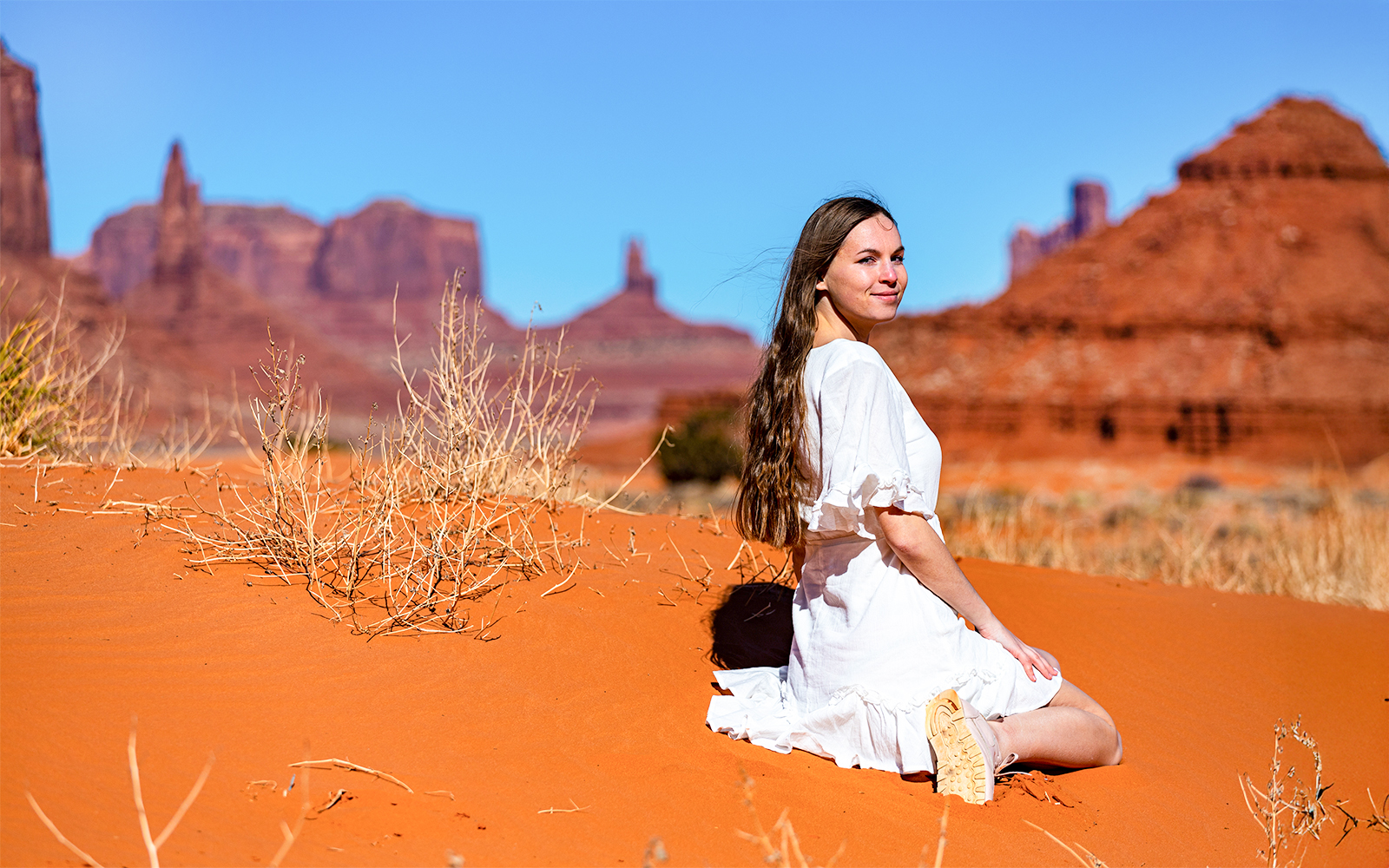 Girl in lace dress sitting on sand with Monument Valley rock formations in background.