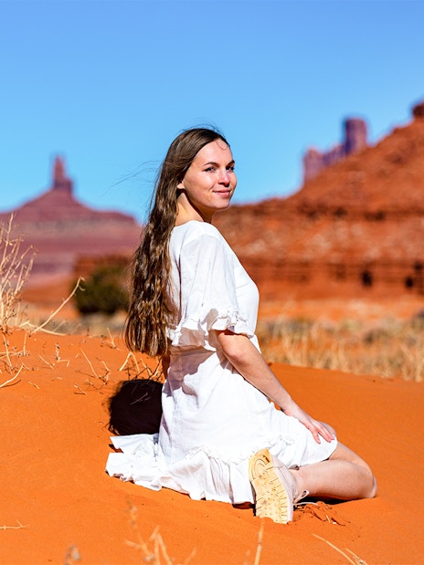 Girl in lace dress sitting on sand with Monument Valley rock formations in background.
