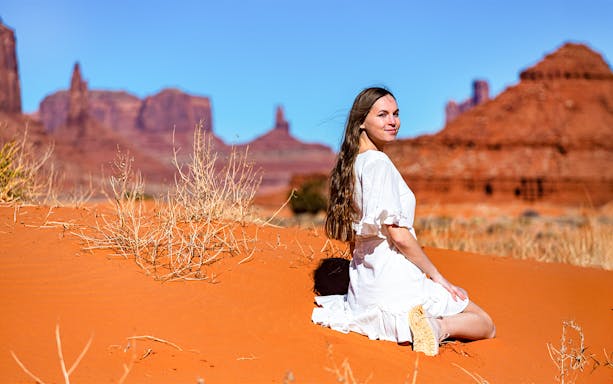 Girl in lace dress sitting on sand with Monument Valley rock formations in background.