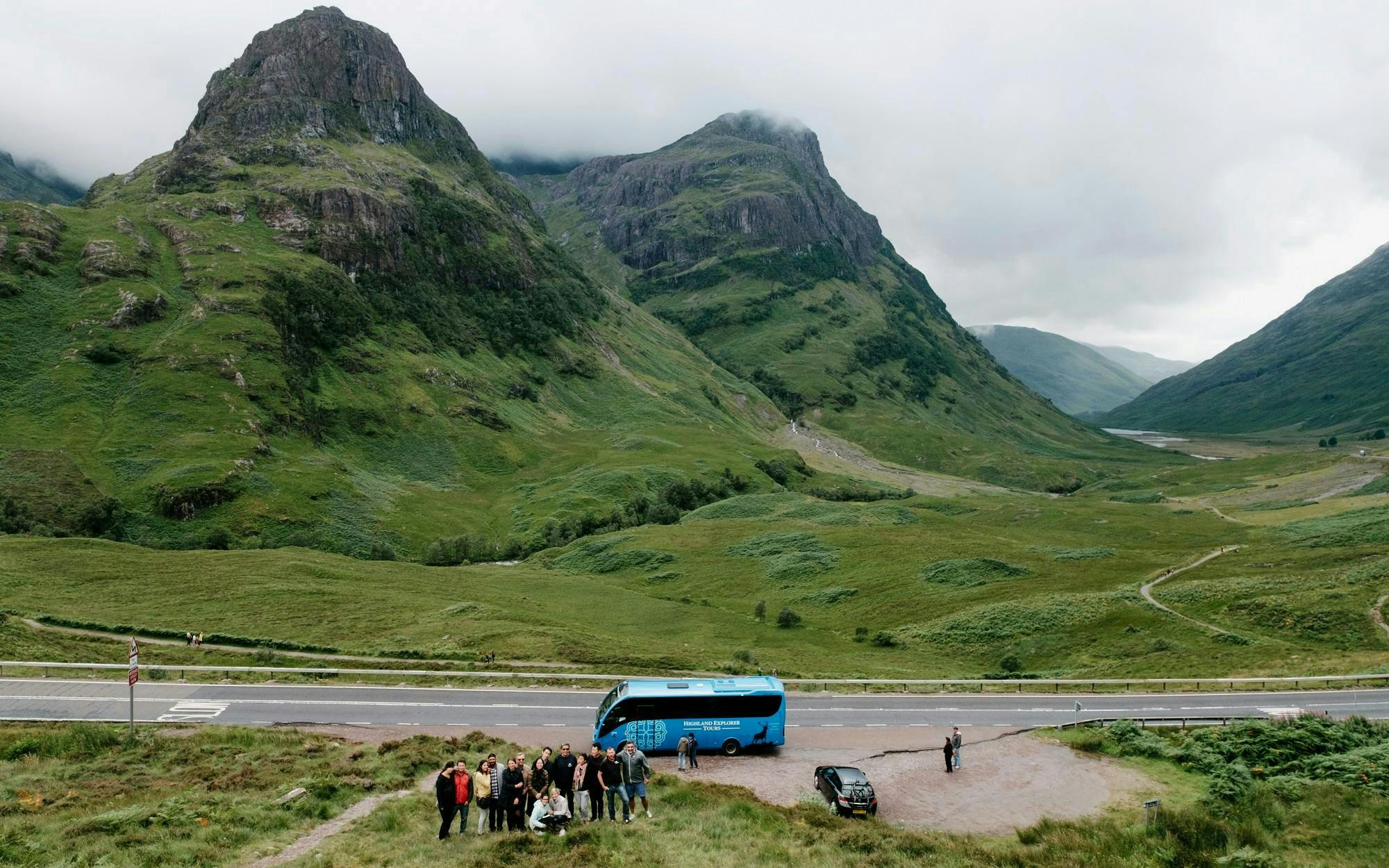Tour group with bus in Glencoe valley, Scotland, surrounded by lush green mountains.