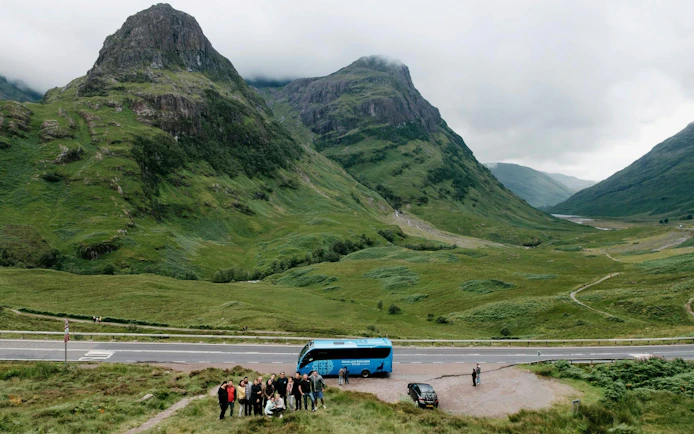 Tour group with bus in Glencoe valley, Scotland, surrounded by lush green mountains.