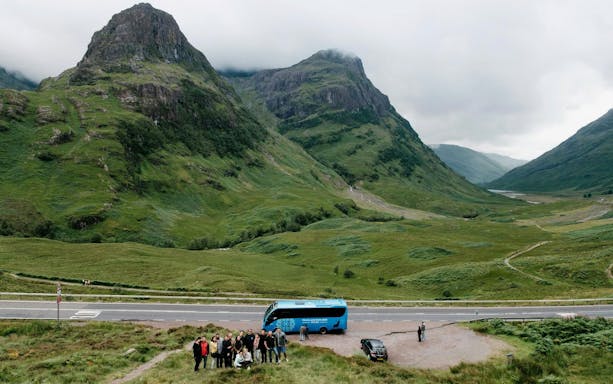 Tour group with bus in Glencoe valley, Scotland, surrounded by lush green mountains.