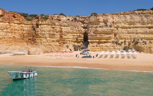Boat near Albufeira beach with umbrellas and BBQ setup against rocky cliffs.