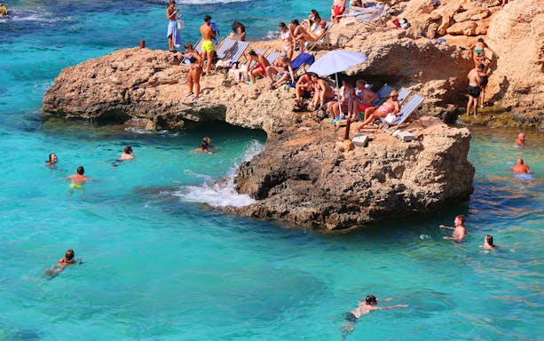 People swimming and relaxing on rocky shore at Blue Lagoon, Malta.