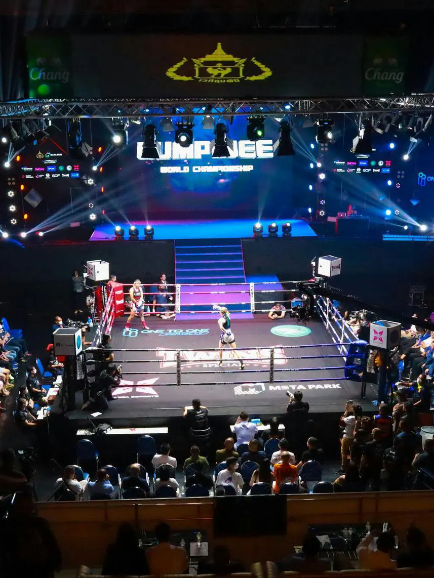 Muay Thai fighters in a match at One Lumpinee Stadium with audience and stage lights.