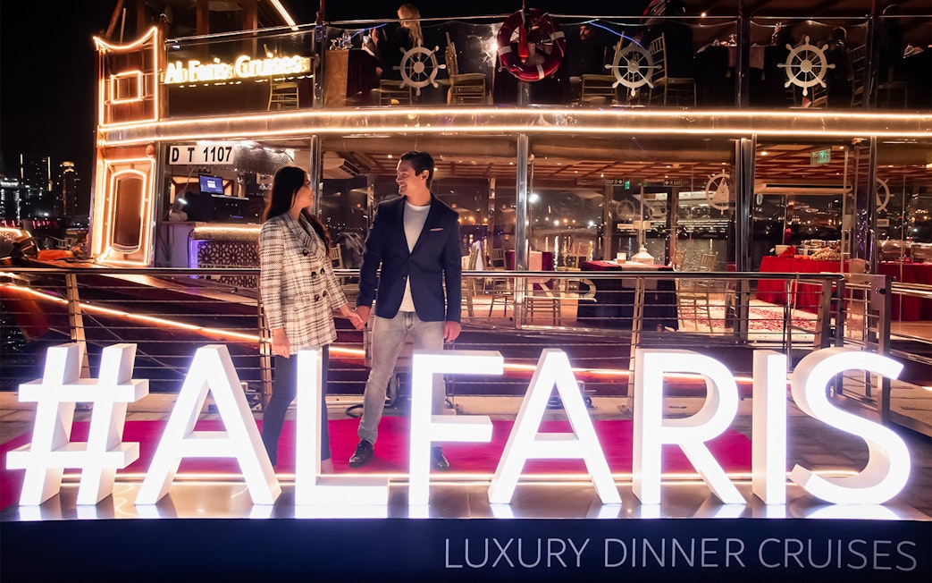 Couple on Dubai Canal Cruise with illuminated Al Faris sign at night.