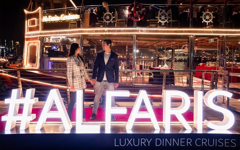 Couple on Dubai Canal Cruise with illuminated Al Faris sign at night.