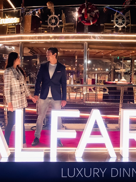 Couple on Dubai Canal Cruise with illuminated Al Faris sign at night.