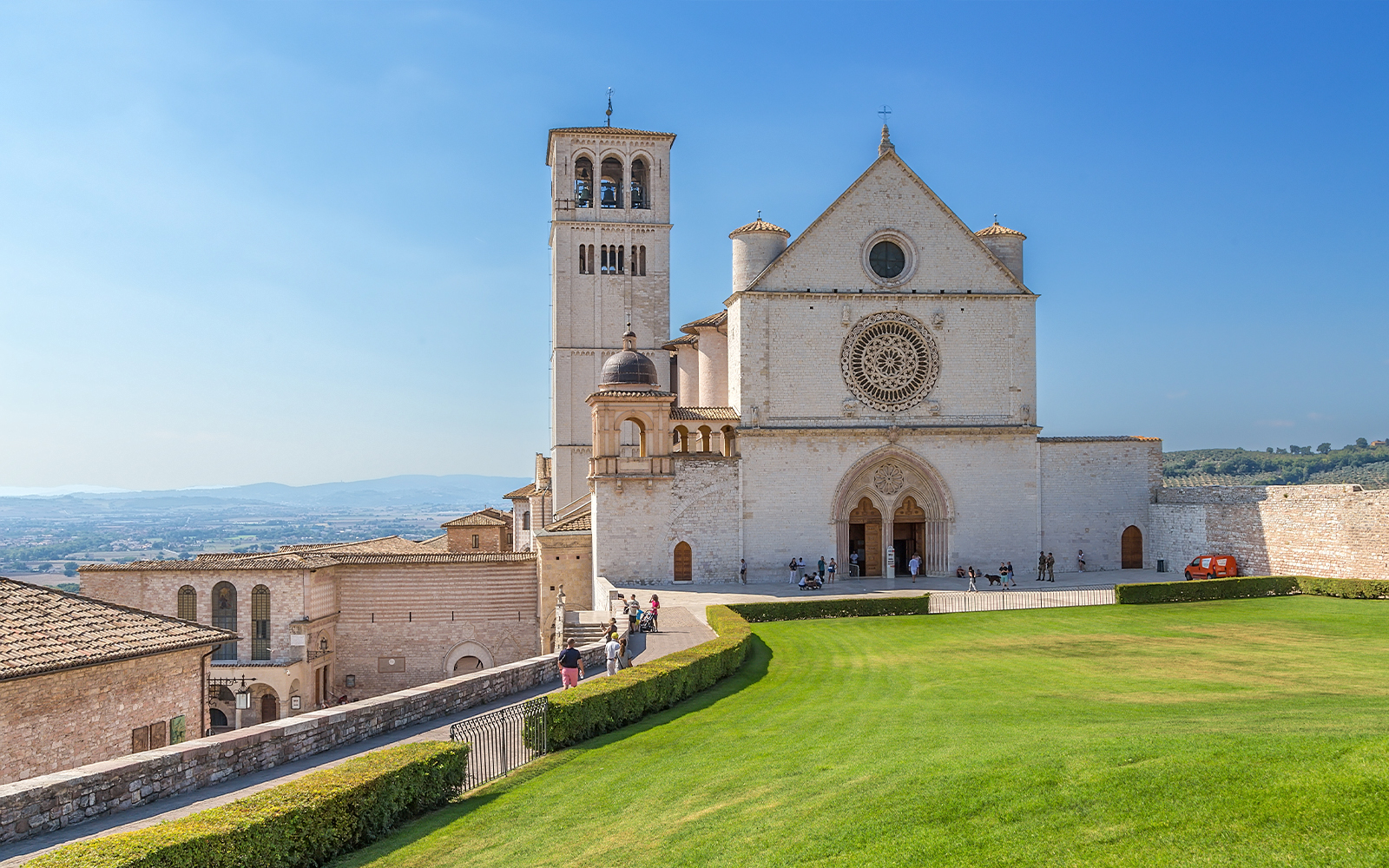 Basilica di San Francesco in Assisi with visitors on the lawn and surrounding architecture.
