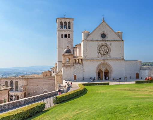 Basilica di San Francesco in Assisi with visitors on the lawn and surrounding architecture.