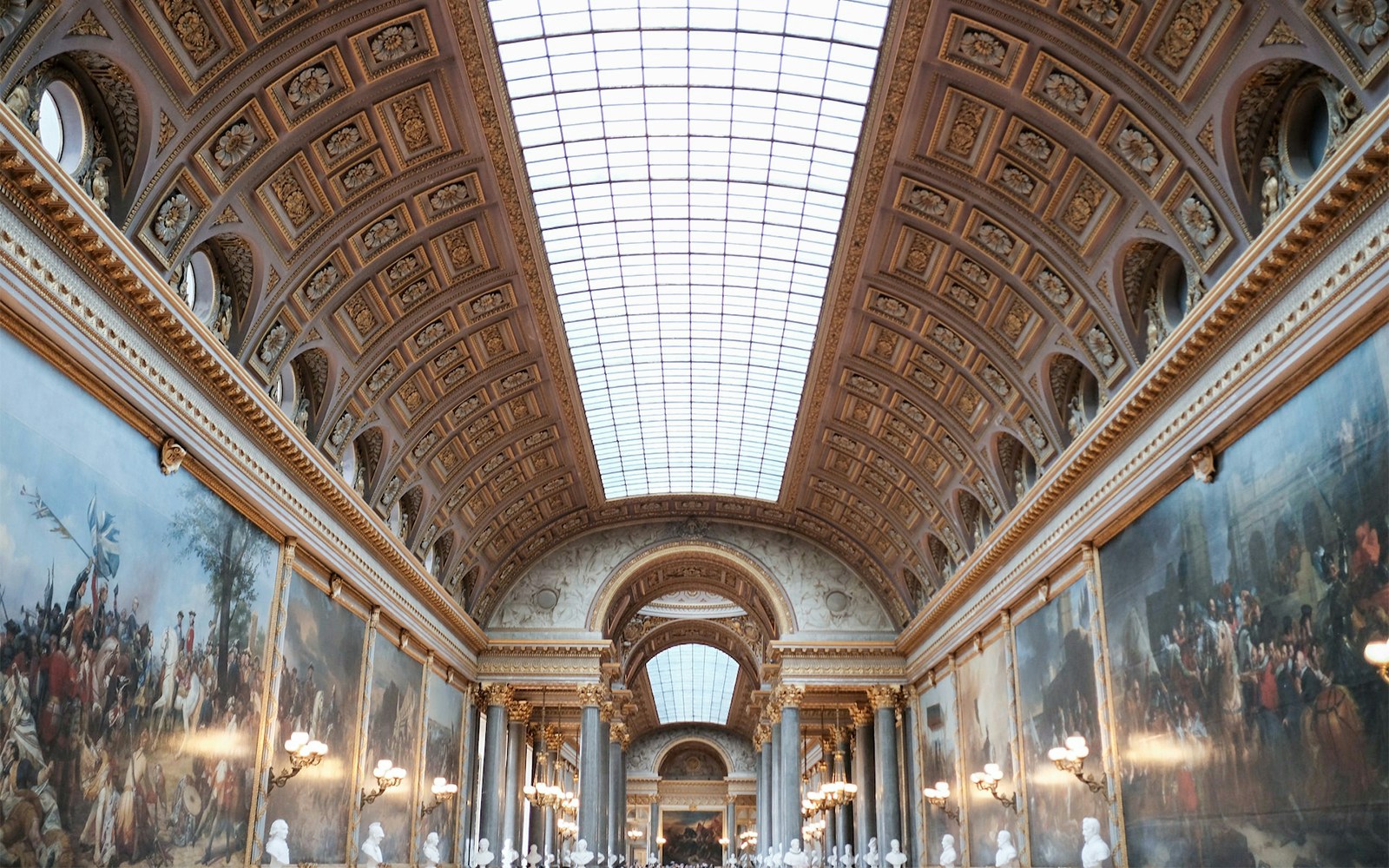 Ein opulenter Raum im Schloss Versailles mit goldenen Verzierungen, einem großen Kamin und Deckengemälden