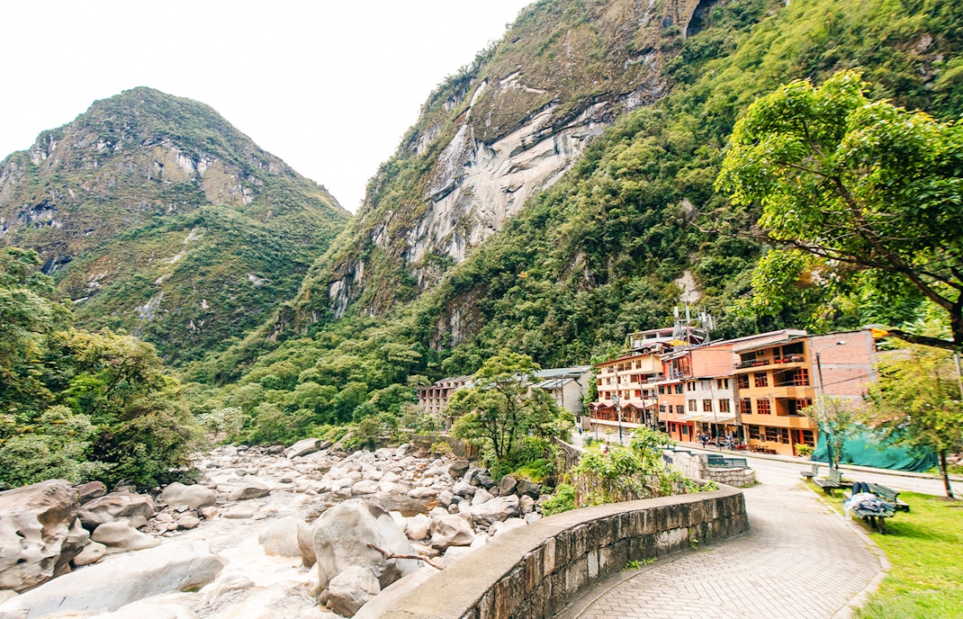 Pathway and buildings along a river in Aguas Calientes, Peru, surrounded by lush mountains.
