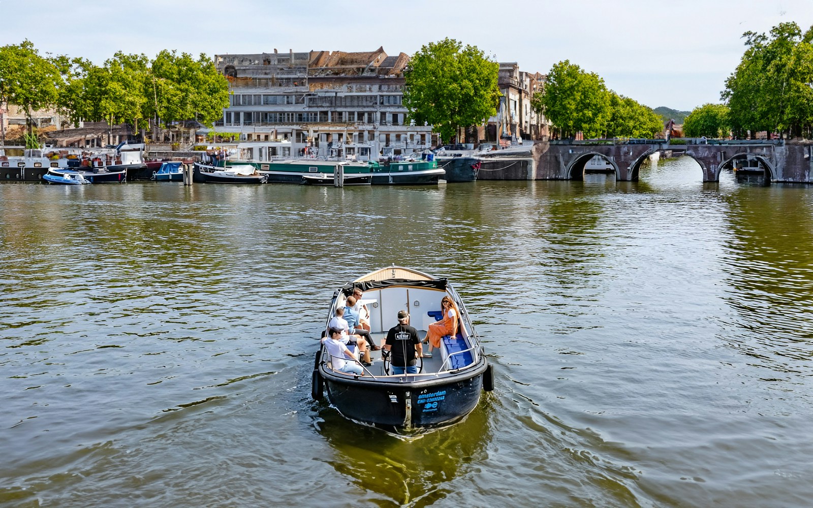 Small open boat cruising Amsterdam canal with passengers, historic buildings, and arched bridge in view.