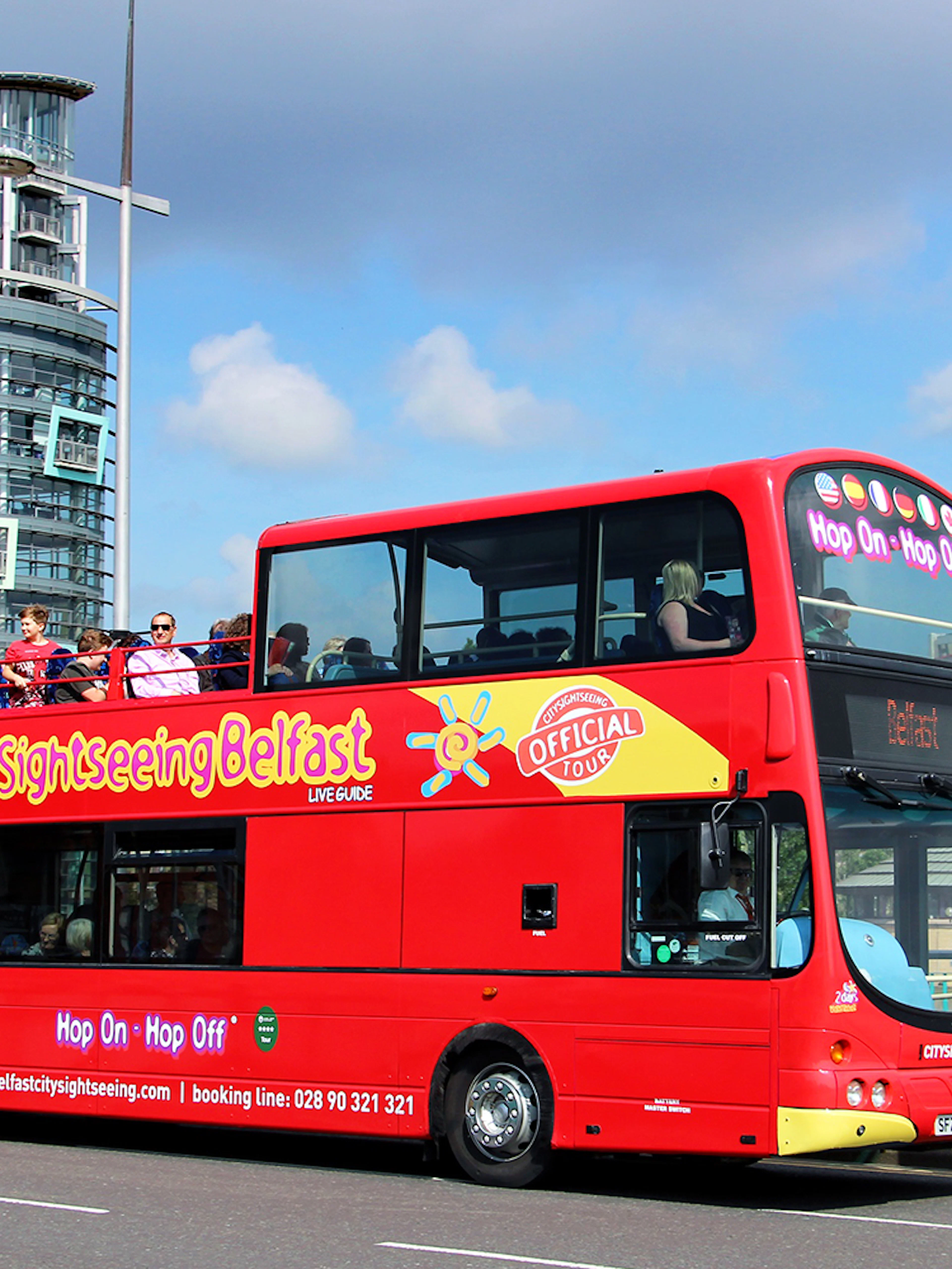 Red double-decker bus on Belfast Hop-On-Hop-Off Tour with city buildings in background.