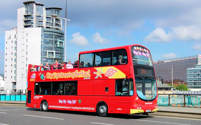 Red double-decker bus on Belfast Hop-On-Hop-Off Tour with city buildings in background.