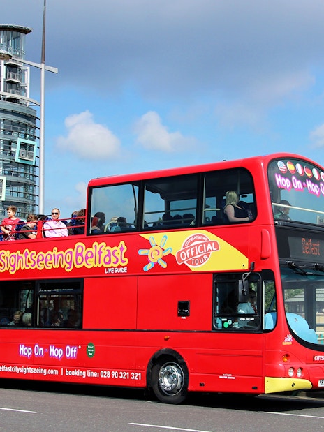 Red double-decker bus on Belfast Hop-On-Hop-Off Tour with city buildings in background.