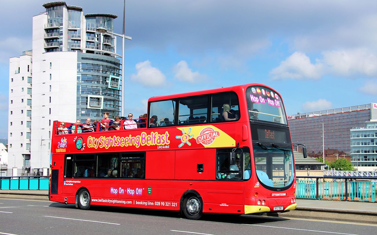 Red double-decker bus on Belfast Hop-On-Hop-Off Tour with city buildings in background.