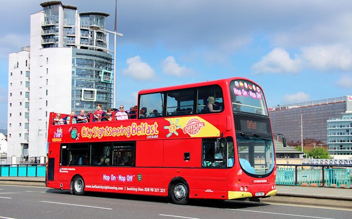 Red double-decker bus on Belfast Hop-On-Hop-Off Tour with city buildings in background.