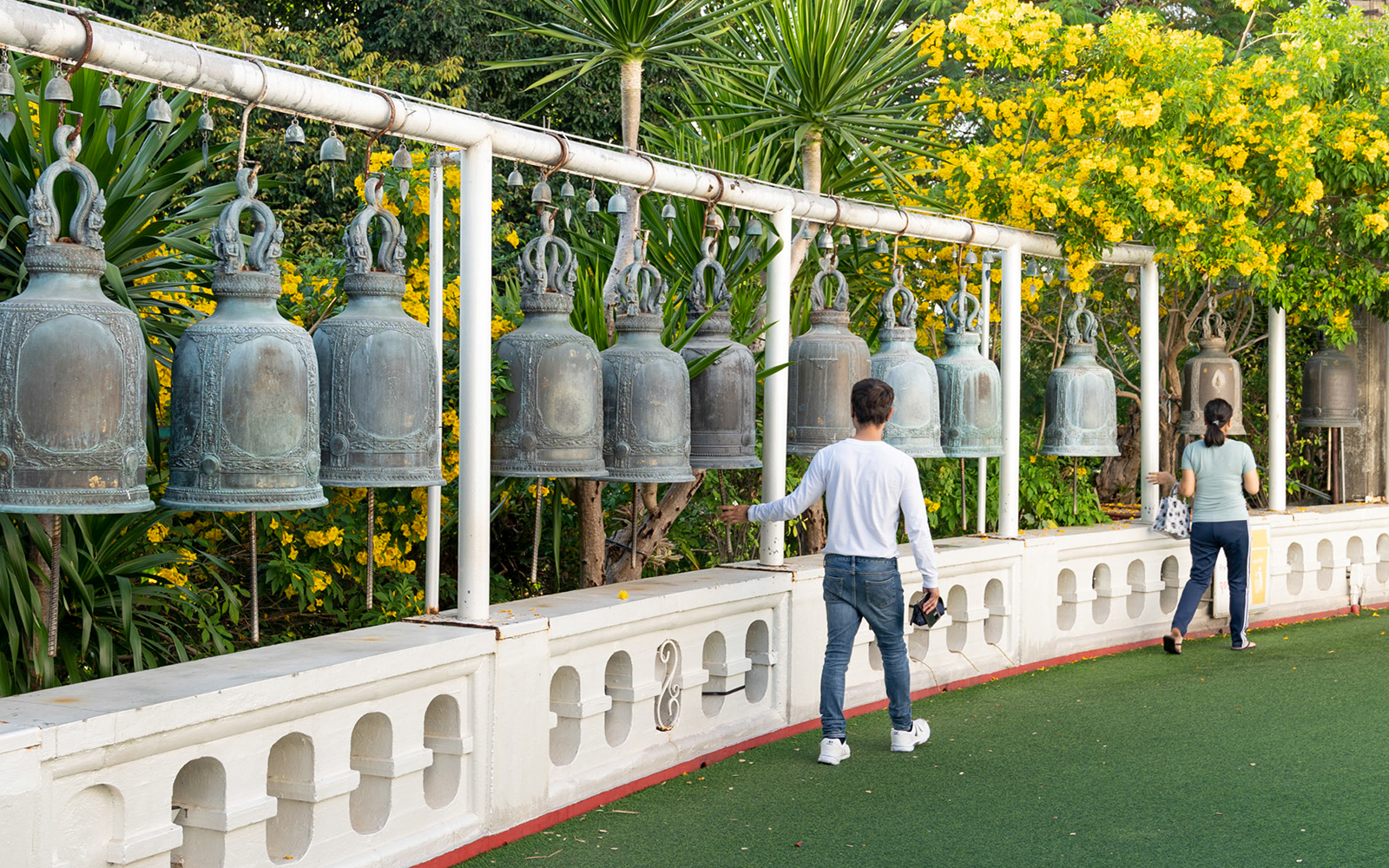 Visitors ringing bells at Wat Pho, Bangkok, surrounded by lush greenery and yellow flowers.