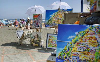 Paintings of Amalfi Coast scenery displayed on a beach with tourists and umbrellas.