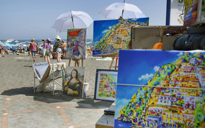 Paintings of Amalfi Coast scenery displayed on a beach with tourists and umbrellas.