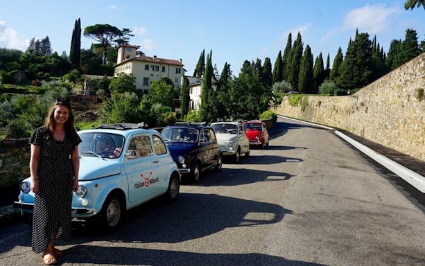Vintage Fiat 500 cars lined up on a scenic road near Florence, Italy, with a participant smiling.