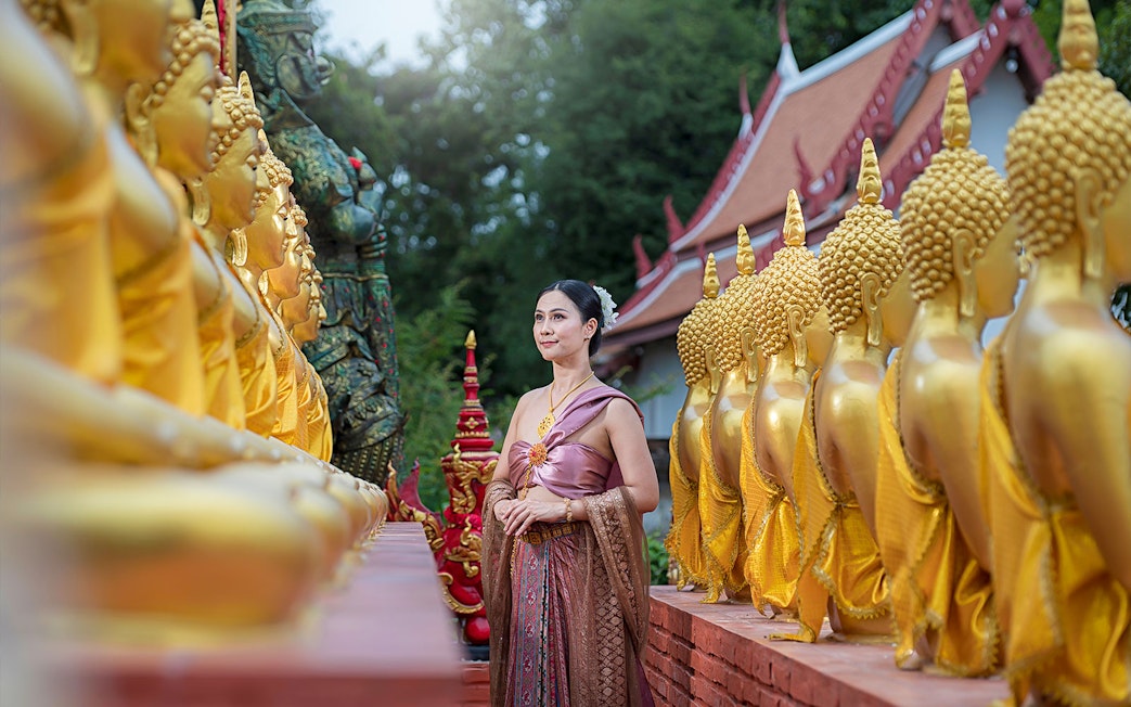 Woman in traditional attire walking between golden Buddha statues at an ancient city temple.