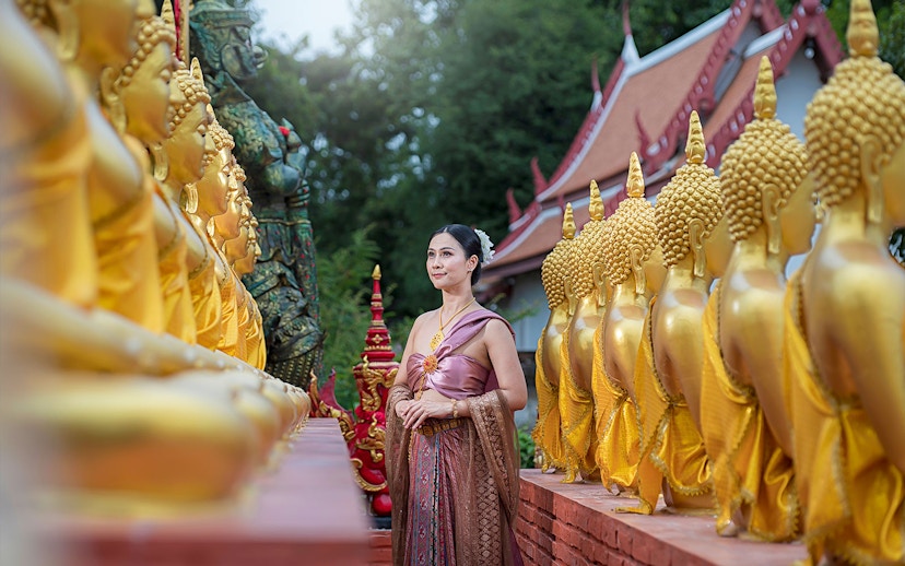 Woman in traditional attire walking between golden Buddha statues at an ancient city temple.