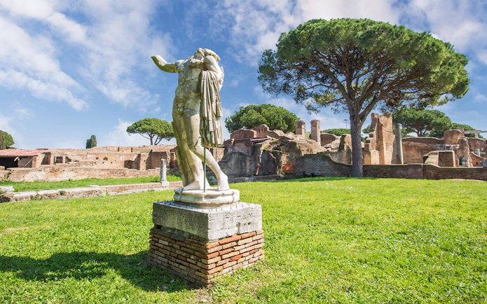 Statue in Ostia Antica ruins with ancient brick structures and trees in the background.