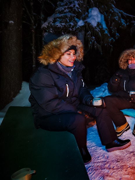 Campers enjoying hot juice by a campfire under Northern Lights in Levi, Finland.