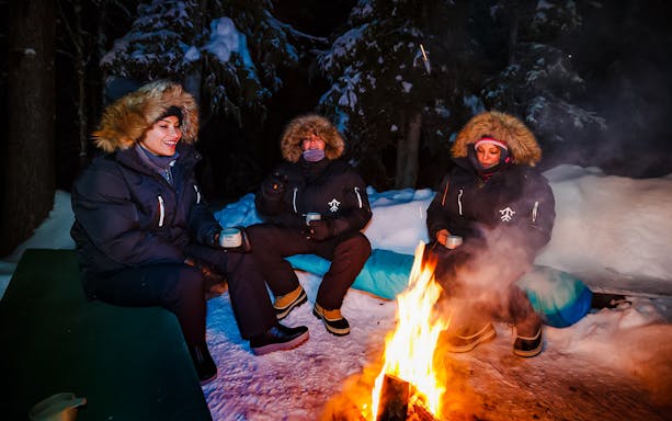 Campers enjoying hot juice by a campfire under Northern Lights in Levi, Finland.