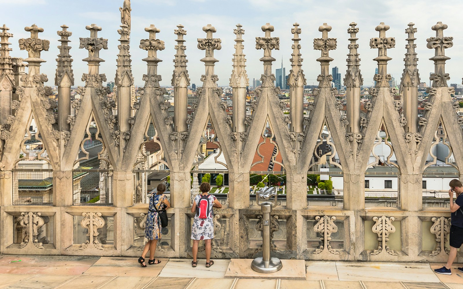 Visitors on the terraces of Milan Cathedral with city view through ornate stonework.