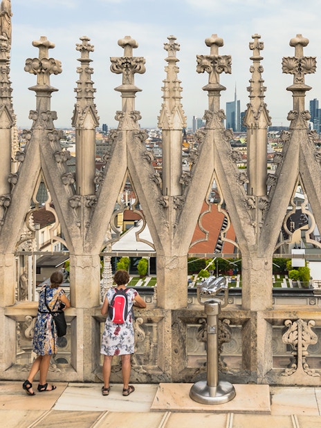Visitors on the terraces of Milan Cathedral with city view through ornate stonework.