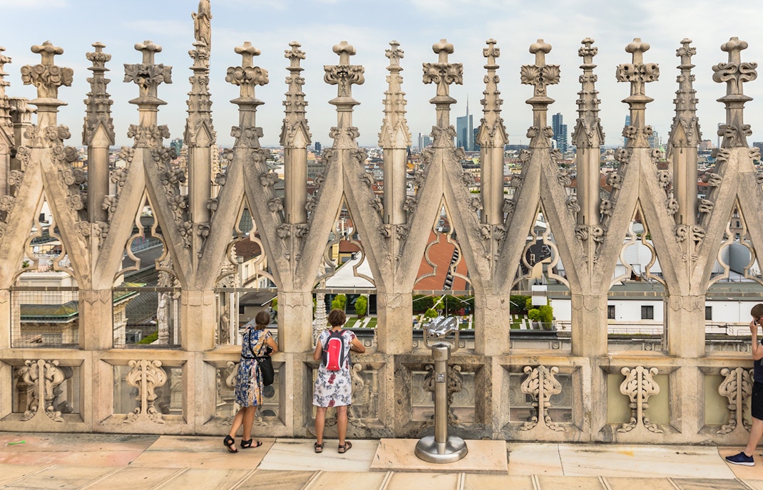 Visitors on the terraces of Milan Cathedral with city view through ornate stonework.
