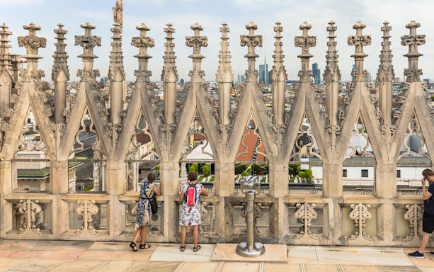 Visitors on the terraces of Milan Cathedral with city view through ornate stonework.