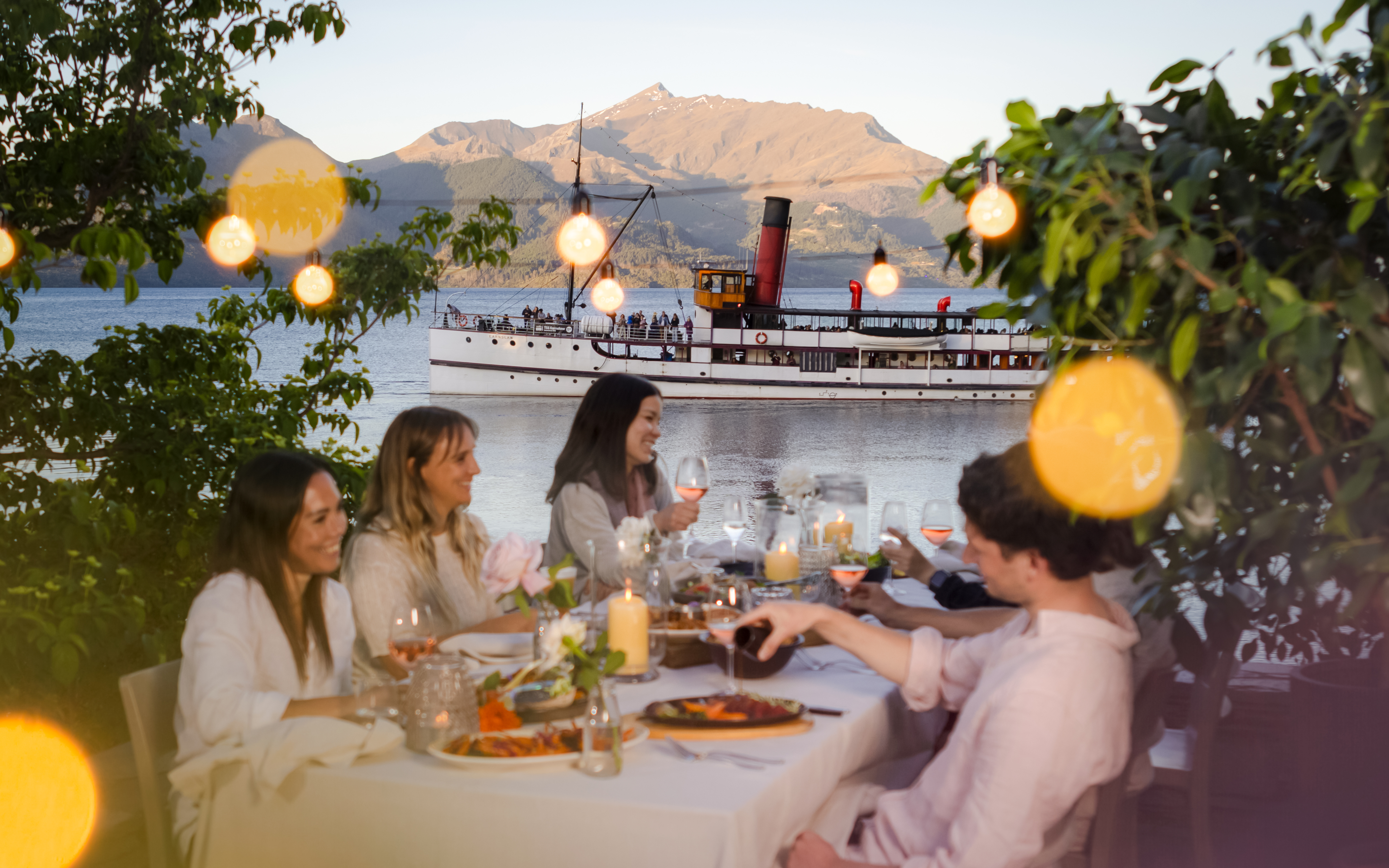 Tourists dining outdoors at Walter Peak High Country Farm with a steamship on Lake Wakatipu.
