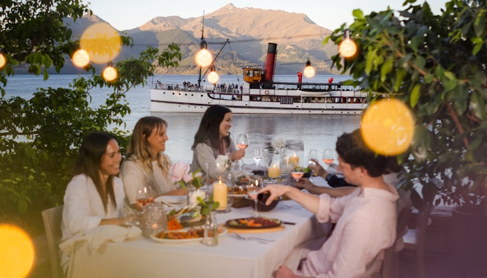 Tourists dining outdoors at Walter Peak High Country Farm with a steamship on Lake Wakatipu.