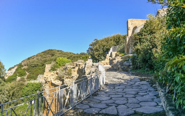 Stone pathway leading through the Archaeological Park of Cumae, surrounded by ancient ruins and greenery.