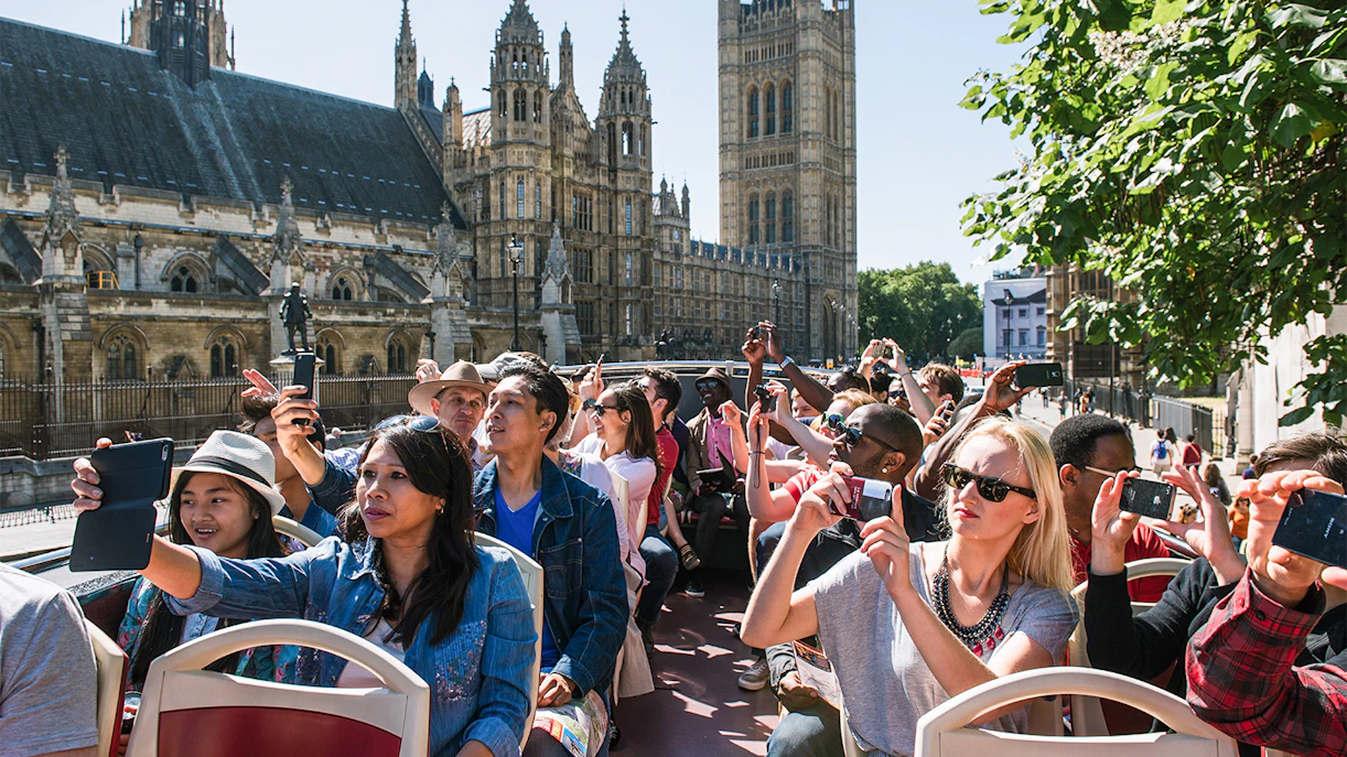 Tourists on an open-top bus tour in London, capturing photos of the Houses of Parliament.