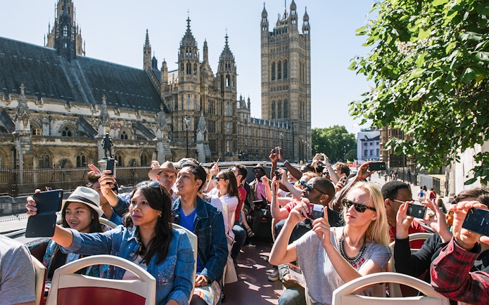 Tourists on an open-top bus tour in London, capturing photos of the Houses of Parliament.