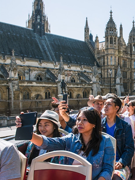 Tourists on an open-top bus tour in London, capturing photos of the Houses of Parliament.