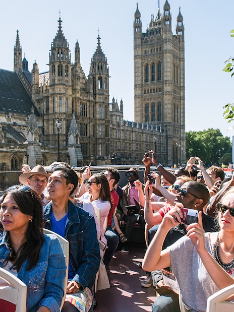 Tourists on an open-top bus tour in London, capturing photos of the Houses of Parliament.