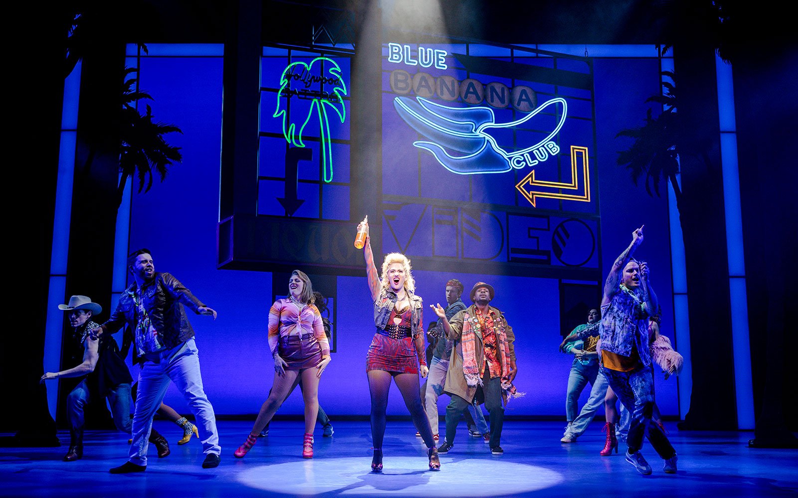 Cast performing a lively scene from Pretty Woman The Musical with colorful neon signs.