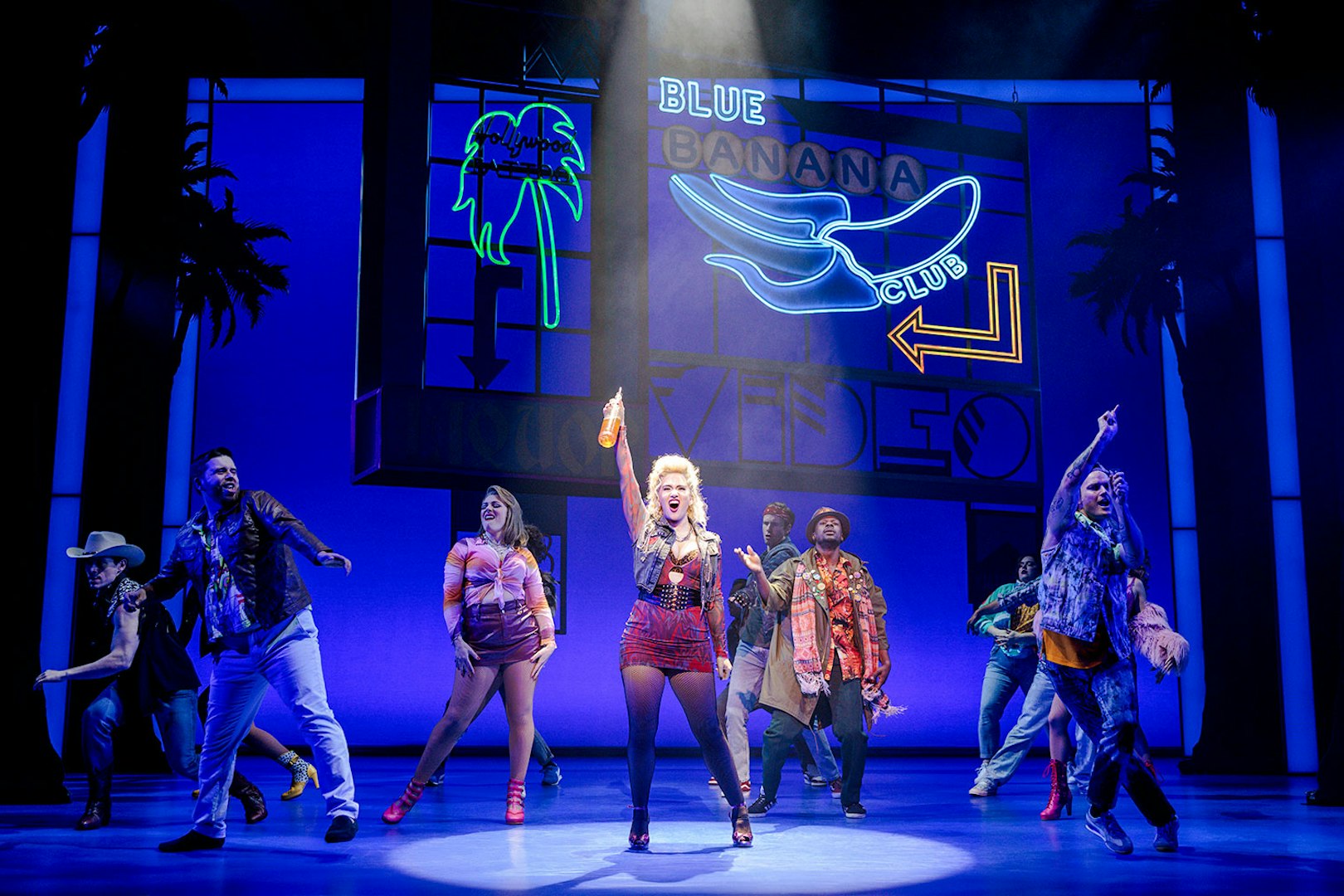 Cast performing a lively scene from Pretty Woman The Musical with colorful neon signs.