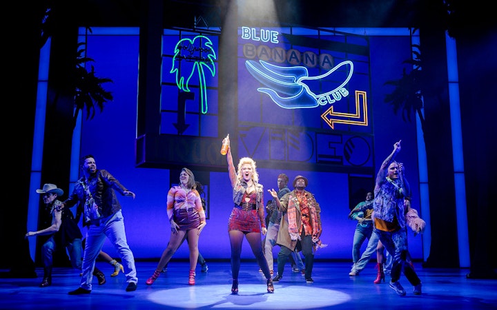 Cast performing a lively scene from Pretty Woman The Musical with colorful neon signs.